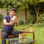 A man sits in a lush green garden, smiling while holding a large jar of Zaiqa Khas pickle. Several other smaller jars are arranged on a glass table in front of him.