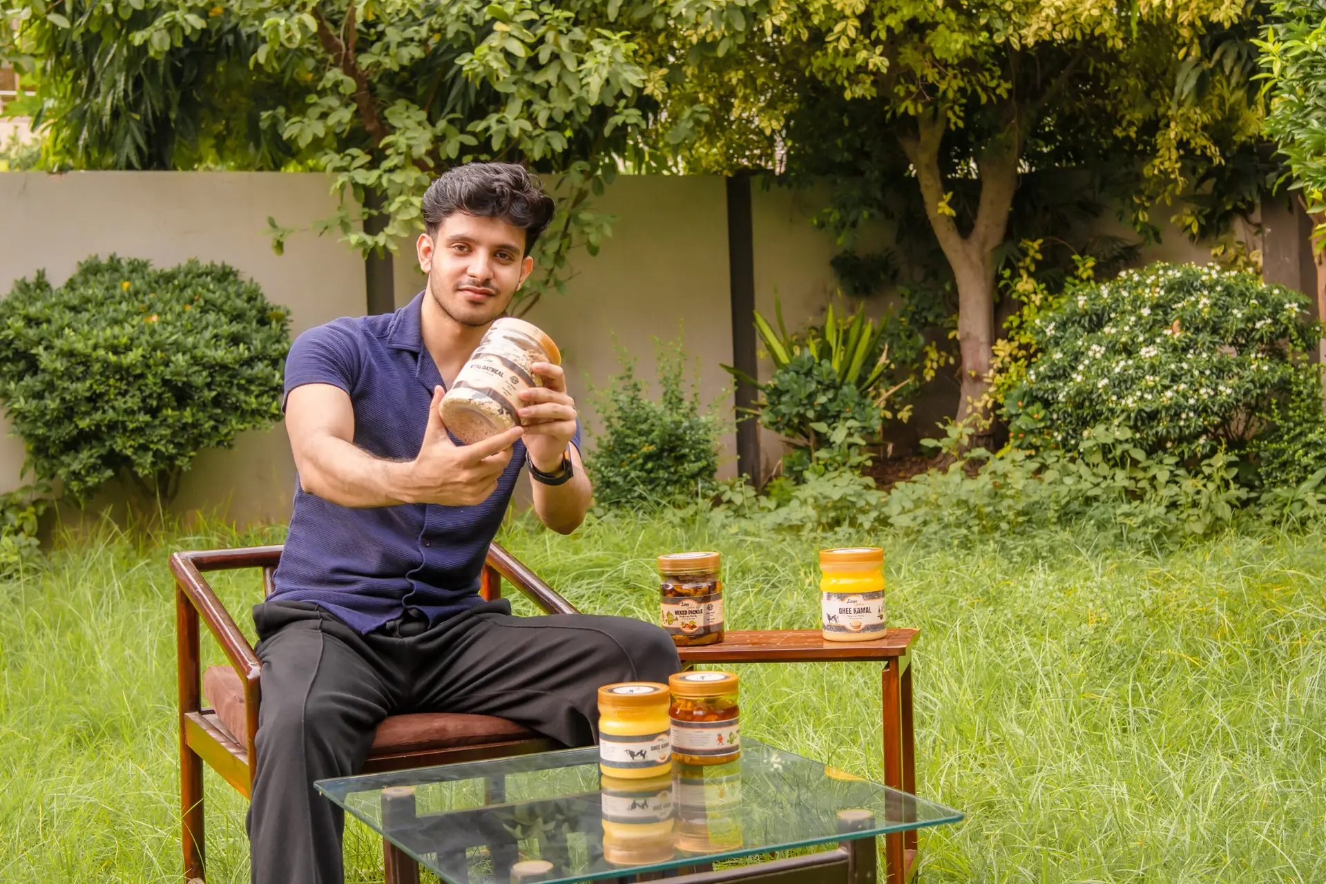 A man sits in a lush green garden, smiling while holding a large jar of Zaiqa Khas pickle. Several other smaller jars are arranged on a glass table in front of him.